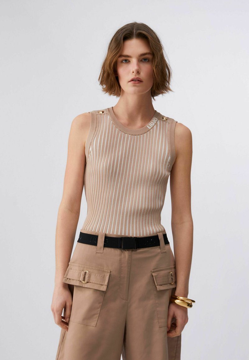 Jeune femme portant un haut sans manches beige et blanc à rayures avec des boutons dorés, un pantalon cargo beige, une ceinture noire et des bracelets dorés.