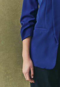 Short-sleeve blue blazer with a smooth texture, side pocket, and a relaxed fit, paired with black pinstriped trousers. Hand resting at side.