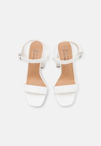 White high-heeled sandals with a glossy finish, featuring two wide straps and an ankle strap secured with a silver buckle. Square toe design.