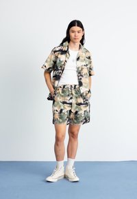 Cămașă cu mânecă scurtă și pantaloni scurți asortați, cu un model de camuflaj în nuanțe de verde, maron și bej. Purtați cu un tricou alb și pantofi sport.