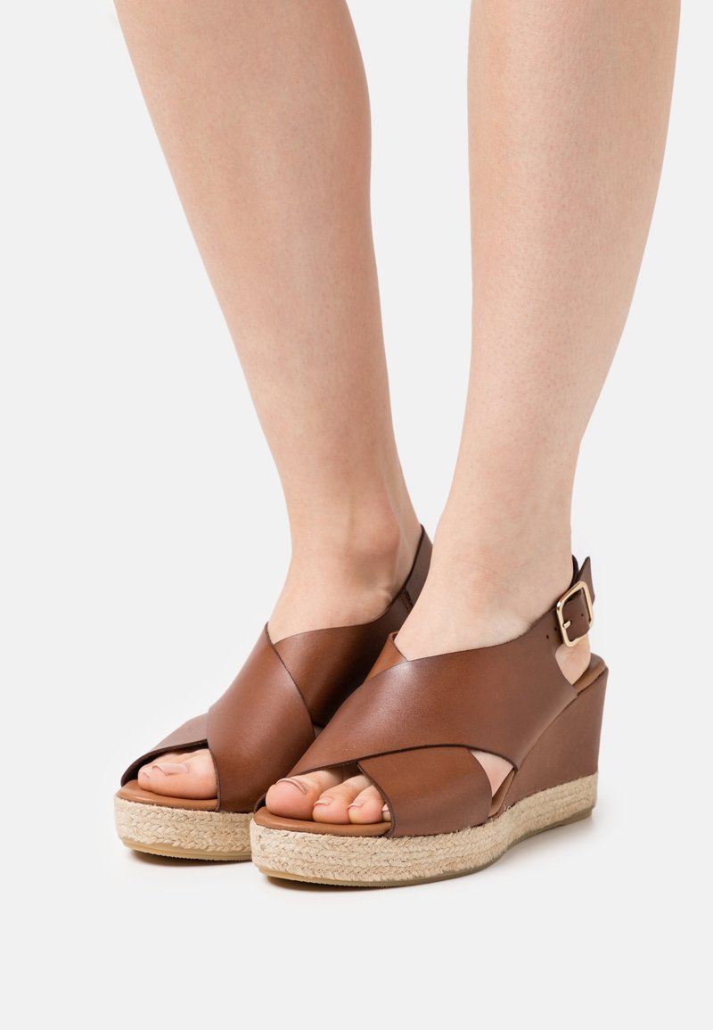 Sandales compensées en cuir brun avec un design à brides croisées, bout ouvert et boucle à la cheville. Texture naturelle de type corde sur la semelle compensée.