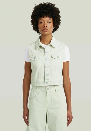Femme aux cheveux bouclés portant un gilet en jean vert clair court sur une chemise blanche à manches courtes et un pantalon taille haute assorti.