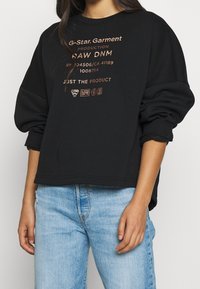 Felpa nera dalla vestibilità ampia, con testo e loghi color rame sul davanti. Tessuto strutturato e polsini a coste completano il design.