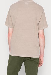 Lyhythihainen, beige T-paita pehmeästä kankaasta. Ominaisuuksiin kuuluu pyöreä kaula-aukko ja näkyvät ompelusaumat helman ja hihansuiden ympärillä.