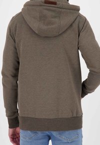 Felpa color verde oliva con cappuccio regolabile, polsini e cintura a costine. Presenta un'etichetta sul retro del collo; il tessuto ha una consistenza liscia.