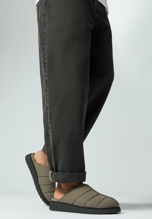 Persona che indossa pantaloni scuri risvoltati e pantofole slip-on imbottite color verde oliva con suole nere, in piedi contro uno sfondo chiaro e uniforme.