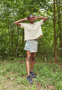 T-shirt oversized beige chiaro, pantaloni corti turchesi con tasca laterale e dettagli riflettenti, scarpe sportive nere in un contesto di foresta verde.