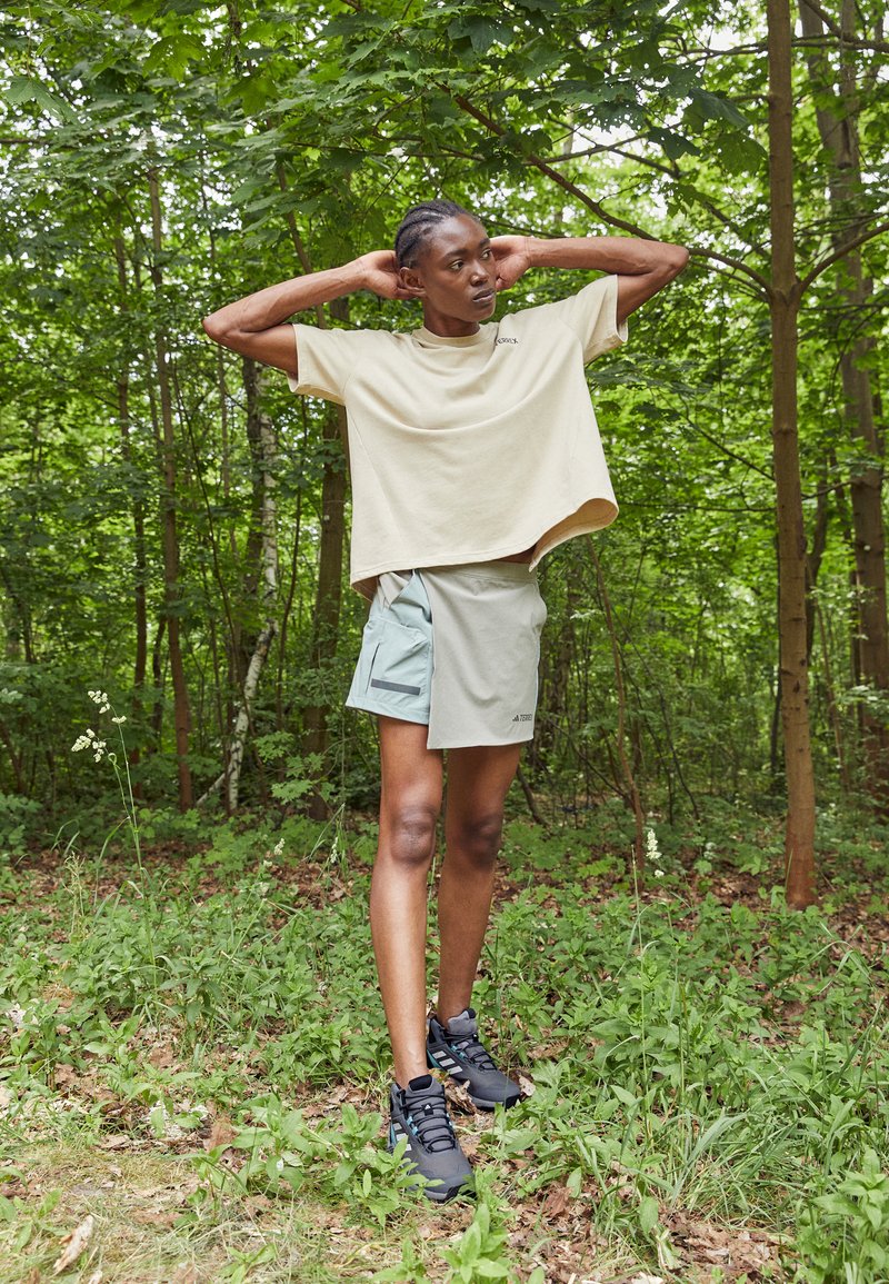 Jasno beżowy, oversize T-shirt, turkusowe szorty z boczną kieszenią i odblaskowym detalem, czarne buty sportowe w zielonym leśnym otoczeniu.