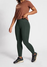 Leggings vert foncé avec une taille haute et une texture côtelée, associés à un t-shirt à manches courtes marron et des baskets noires avec un accent multicolore.