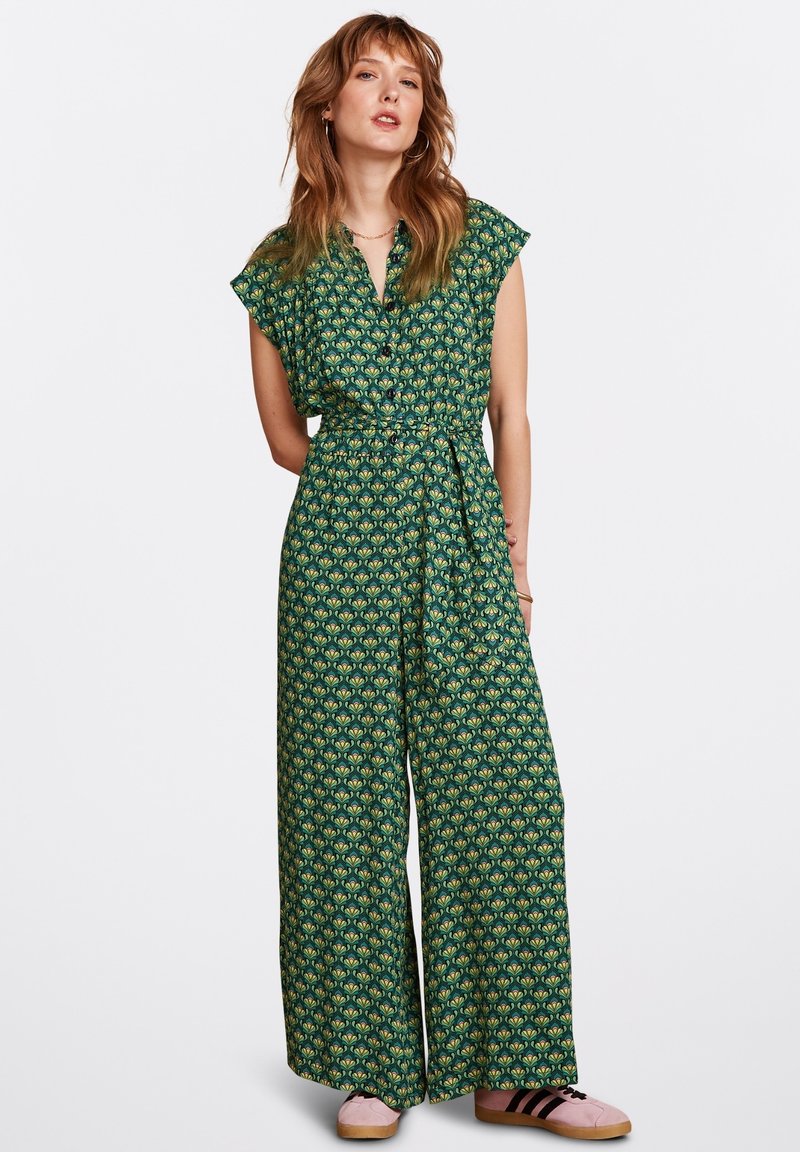 Grön blommig jumpsuit med korta ärmar, markerad midja, vida ben och ett upprepat bladmönster. Bärs med rosa sneakers.