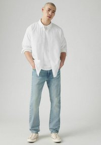 Weißes Hemd mit Knöpfen, darunter ein weißes T-Shirt, helle blaue Jeans und beigen Sneakern. Glatter Stoff, lässiger Schnitt, lässiger Stil.