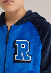 Blå och marinblå hoodie i fleece med dragkedja, med en framträdande broderad bokstav "R" i blått och vitt på bröstet.