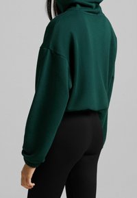 Femme portant un haut à col roulé à manches longues vert foncé et un pantalon noir ajusté à côtes, vue de dos sur un fond gris uni.