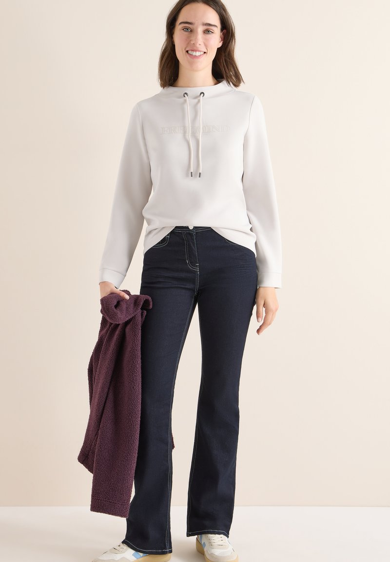 Lichtgrijze hoodie met de tekst "FREE MIND", gecombineerd met donkerblauwe flare jeans en een bordeaux fleece in de hand. Witte sneakers maken de look af.