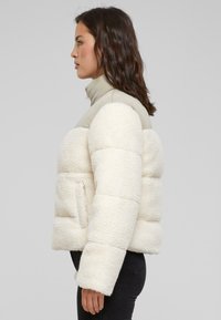Veste rembourrée couleur crème avec une texture en fausse fourrure, col montant et deux poches avant. Présente une partie supérieure lisse contrastante.