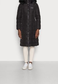 Giacca a vento nera con cappuccio in pelliccia sintetica, chiusura con zip e motivo trapuntato verticale. Indossata sopra leggings crema e sneakers bianche.