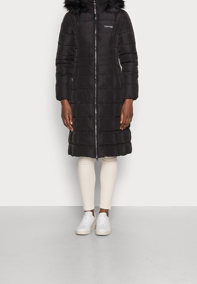 Giacca a vento nera con cappuccio in pelliccia sintetica, chiusura con zip e motivo trapuntato verticale. Indossata sopra leggings crema e sneakers bianche.