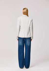 Blazer gris clair sur mesure avec épaules structurées, associé à un jean bleu à taille haute et jambes larges, doté de poches arrière et d'une finition délavée.