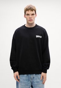 Felpa nera realizzata in morbido tessuto con scollo rotondo, spalle scese e un logo bianco "Hugo" sul lato sinistro del petto. Abbinata a jeans blu chiaro.