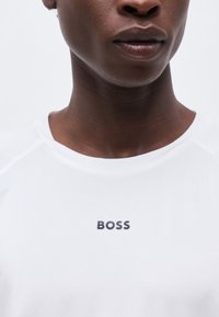 Κοντινό πλάνο ενός ατόμου που φοράει λευκό πουκάμισο με την επιγραφή «BOSS» σε μαύρα γράμματα στο στήθος, μπροστά σε απλό φόντο.