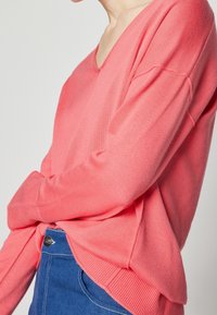Suéter de punto coral con escote en V, hombros caídos y puños de canalé, combinado con pantalones de mezclilla azules con un botón plateado.