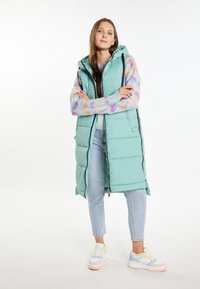 Gilet imbottito lungo verde menta con zip, dotato di cappuccio e tasche laterali. Indossato sopra un maglione a motivi multicolori e jeans chiari.