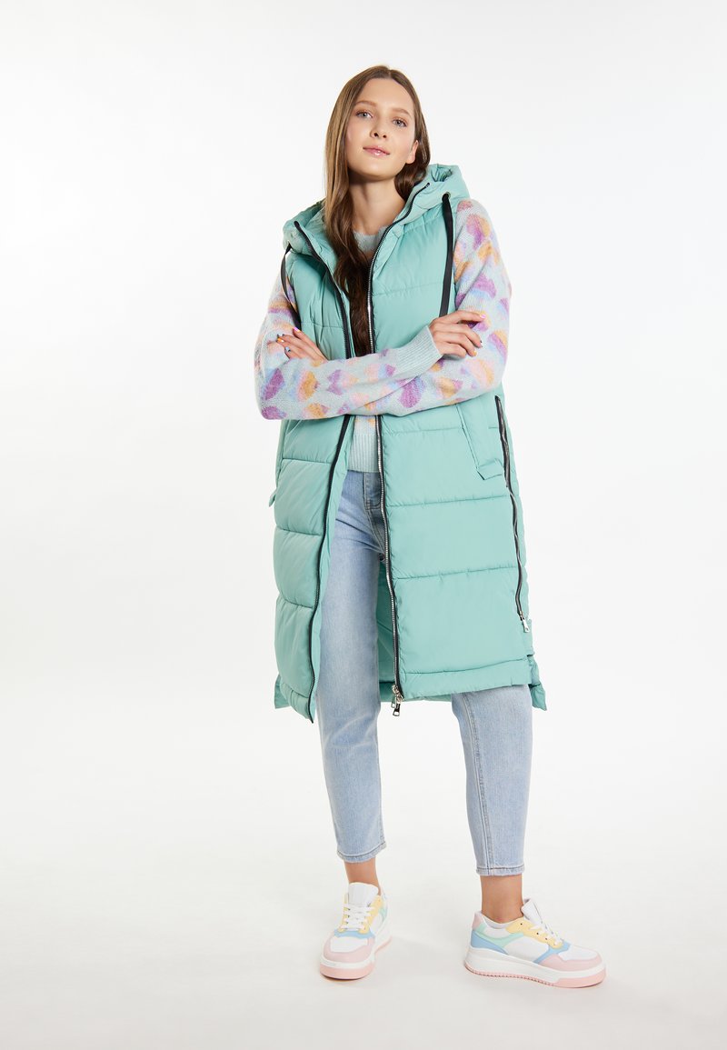 Gilet imbottito lungo verde menta con zip, dotato di cappuccio e tasche laterali. Indossato sopra un maglione a motivi multicolori e jeans chiari.