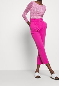 Vitt, figursydd långärmad topp i rosa med blommönster, matchad med högmidjade rosa byxor med bälte och avklippta, smalnande ben. Vita slidesandaler.