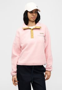 Φλις πουλόβερ σε ανοιχτό ροζ χρώμα με κίτρινο κλείσιμο με κουμπιά, φερμουάρ στο στήθος και λογότυπο. Διαθέτει μακριά μανίκια και άνετη γραμμή.