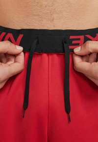 Mani che regolano il cordoncino nero dei pantaloni sportivi rossi con cintura nera, presentante scritte rosse, su un torso maschile.