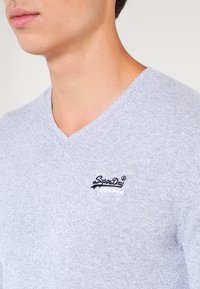 Jasnoszary sweter z dekoltem w serek, wykonany z teksturowanej tkaniny, z małym granatowym wyszywanym logo na lewej piersi.