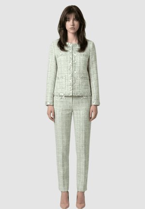 Mujer que lleva una chaqueta de tweed verde pálido con botones y pantalones de talle alto a juego, de pie frente a un fondo claro y liso.