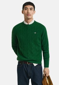 Groene kabelgebreide trui met ronde hals, geribbelde manchetten en subtiele logo-detail. Gecombineerd met donkere jeans en een bruine tas in de hand.