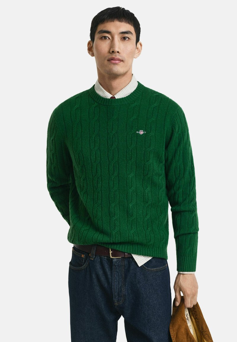 Groene kabelgebreide trui met ronde hals, geribbelde manchetten en subtiele logo-detail. Gecombineerd met donkere jeans en een bruine tas in de hand.