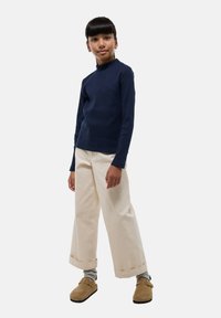 Haut à manches longues côtelé marine, pantalon large crème, chaussettes grises et sabots marron clair. Design simple et fonctionnel avec des poignets retroussés.