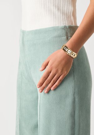 Gold bracelet with square face, paired with light green corduroy pants featuring vertical ribbed texture. Hand resting on pants.