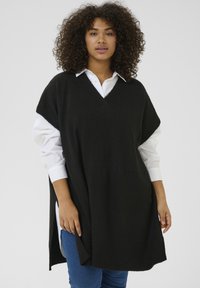 Czarny oversize sweter w formie ponczo z dekoltem w serek, z bocznymi rozcięciami i ściągaczem na dole. Noszony na białej koszuli z guzikami i niebieskich dżinsach.