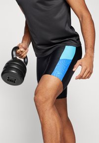 Fekete sportnadrág kék mintázatú oldalsó panelekből, egy személy viseli, aki egy fekete kettlebellt tart, sötétszürke pólóval párosítva.