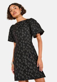 Schwarzes Kleid mit strukturiertem floralen Stickerei, Puffärmeln und einer taillierten Silhouette mit ausgestelltem Saum. Glatter Stoff mit bemerkenswerten Details.