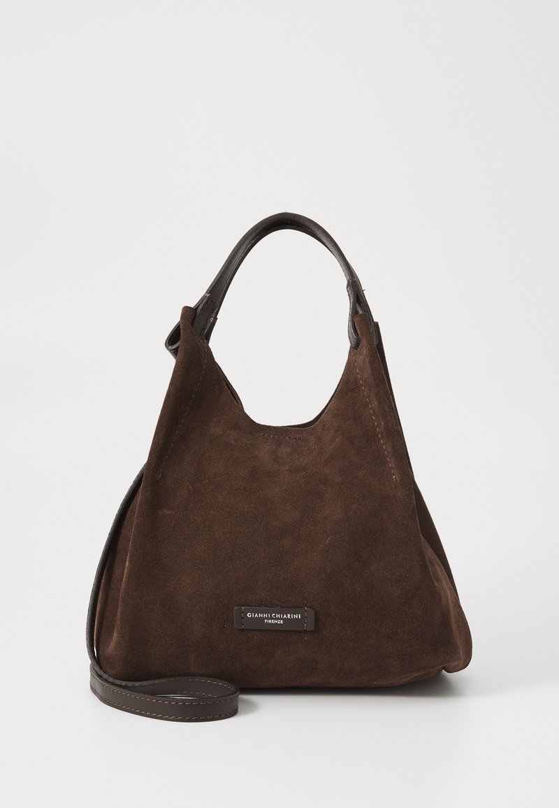Bruine suède handtas met een zachte textuur, met een gebogen silhouet, korte leren handvatten en een logo plaatje nabij de basis.