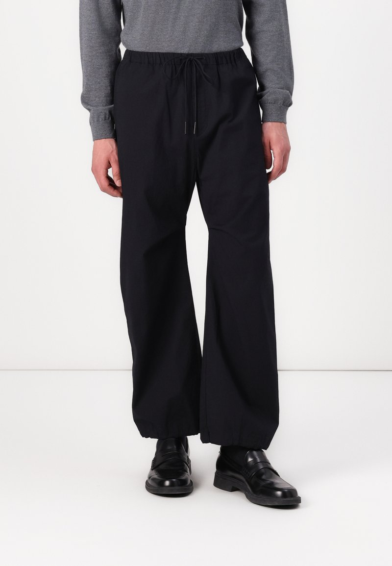 Pantalons larges noirs avec une taille élastique et un cordon de serrage. Texture du tissu lisse. Portés avec des chaussures en cuir noires.