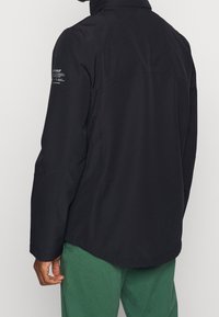 Černá větrovka s vysokým límcem, hladkým povrchem a logem na levém rukávu. Má elegantní design a moderní střih.