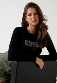 Zwarte trui met "GUESS" in strass, ronde halslijn, lange mouwen, gecombineerd met olijfgroene shorts, zittend tegen een zwarte stoel.