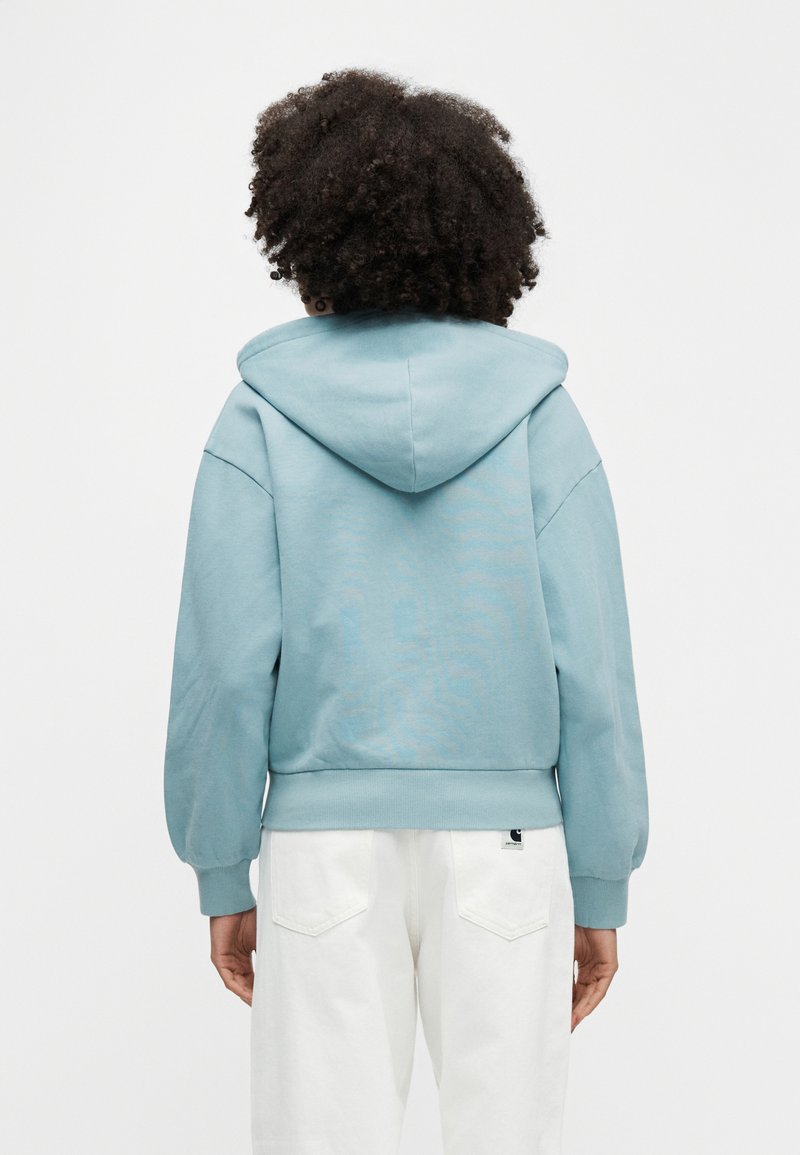 Personne aux cheveux bouclés portant un sweat à capuche bleu clair et un pantalon blanc, vue de dos sur un fond uni.