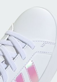 Bílá teniska s hladkým syntetickým svrškem, texturovanými bílými tkaničkami a duhovými růžovými pruhy na straně. Zaoblená špička a minimální prošití.