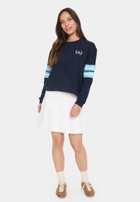 Felpa navy con strisce azzurro chiaro sulle maniche, scollo rotondo e logo. Abbinata a una gonna bianca plissettata e sneakers marroni.