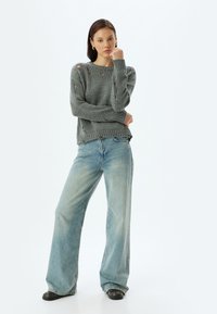 Grijze gebreide trui met geribbelde manchetten, voorzien van versleten gaten. Gecombineerd met lichtblauwe wijde jeans en zwarte enkellaarsjes.