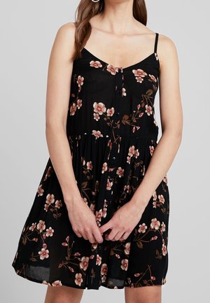 Kobieta w czarnej, bezrękawnikowej sukience zapinanej na guziki z różowo-brązowym kwiatowym nadrukiem, ręce delikatnie złożone z przodu, neutralne tło.