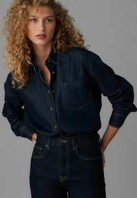 Dunkelblaue Jeanshemd mit langen Ärmeln, Knopfverschluss und einer Brusttasche. Kombiniert mit passenden dunklen Jeans, die kontrastierende Nähte aufweisen.