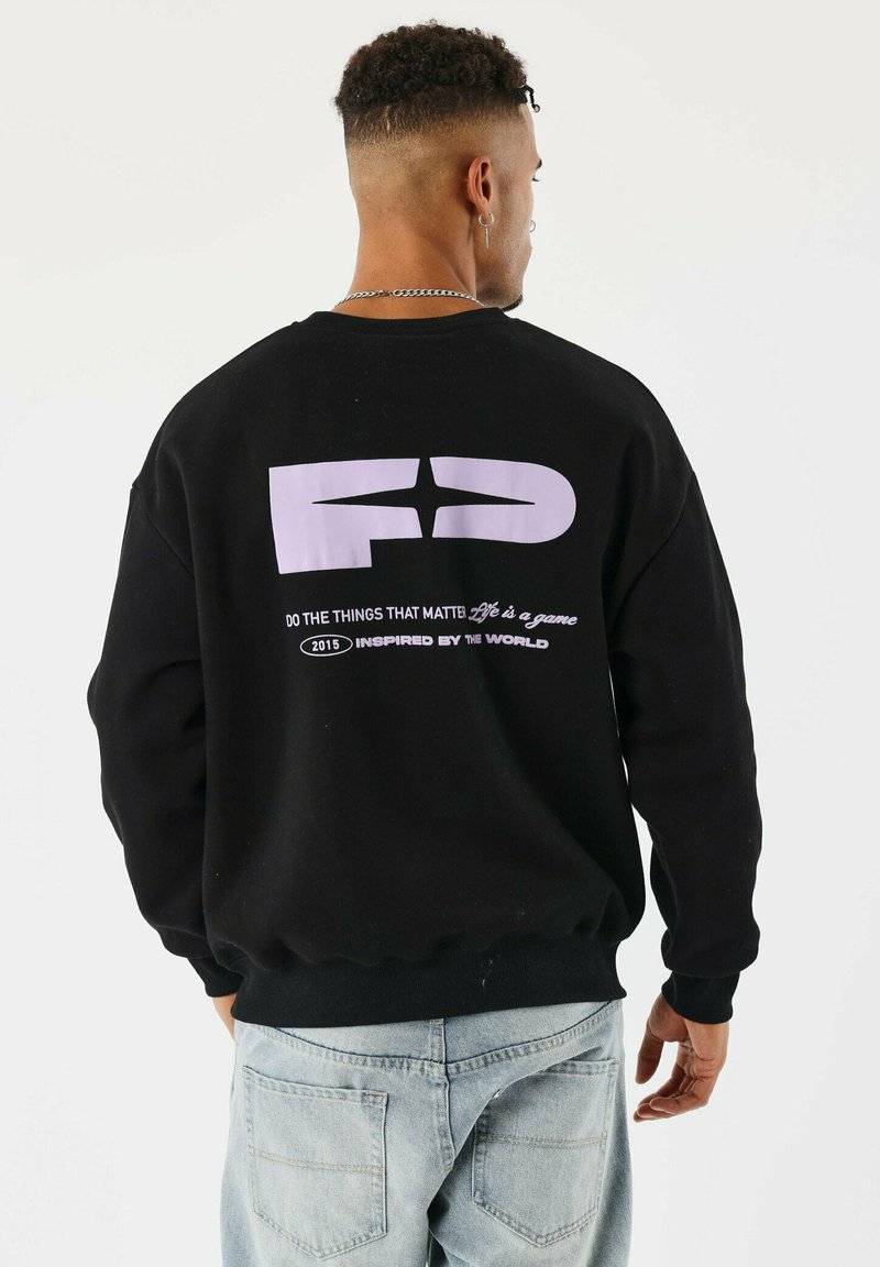 Homme portant un sweat-shirt noir avec un graphisme violet et le texte "Faites les choses qui comptent, la vie est un jeu, 2015 inspiré par le monde" au dos.